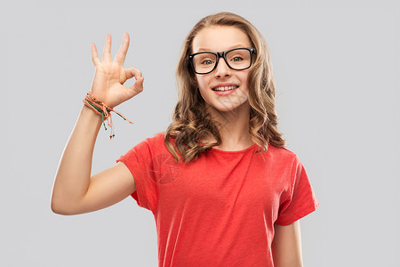 好视力字体设计教育,学校人的微笑的十几岁的学生女孩戴着眼镜红色T恤,灰色背景上OK手牌微笑的学生女孩戴着眼镜,出良好的背景