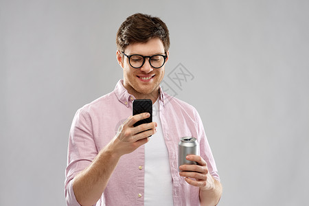 锡罐电话技术,互联网人的微笑的轻人戴着眼镜,看着智能手机,喝着锡罐里的苏打水,背景灰色的戴眼镜的轻人带着智能手机饮料背景