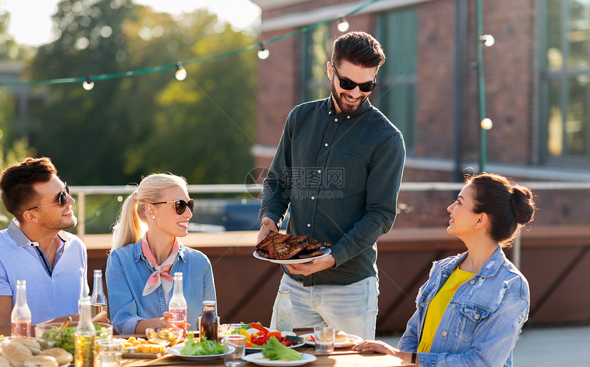 休闲人们的快乐的聚会主持人提供肉给他的朋友烧烤屋顶夏天夏天屋顶上的烧烤派上的朋友图片