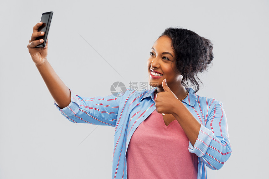 技术手势人们的快乐的非裔美国妇女用智能手机自拍,并灰色背景上竖大拇指非裔美国妇女用智能手机自拍图片