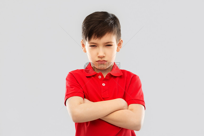 童,表情人的高兴的小男孩交叉手臂红色马球T恤撅嘴眯眼灰色的背景穿着t恤的高兴的男孩撅着嘴眯着眼睛图片