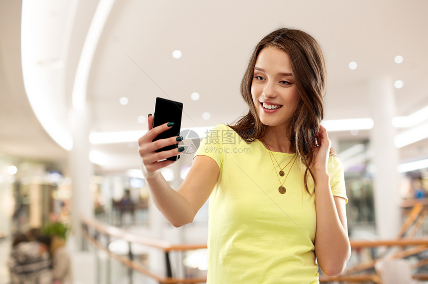 技术人的微笑的轻妇女十几岁的女孩穿着空白的黄色T恤,购物中心背景下智能手机自拍十几岁的女孩购物中心自拍图片