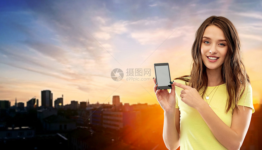 技术人的微笑的轻妇女十几岁的女孩T恤指着智能手机空白屏幕日落塔林城市背景持智能手机的轻女少女图片