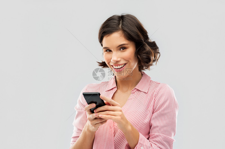 技术人的快乐的微笑轻妇女穿着条纹衬衫用智能手机灰色背景穿着条纹衬衫的轻女人用智能手机图片