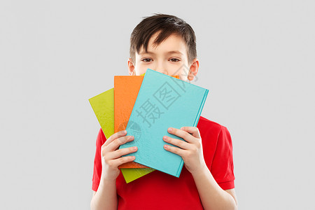 学校,教育人的小学生男孩穿着红色T恤,躲灰色背景的书后害羞的小学生躲书后图片