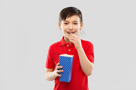 小男孩抬着水桶快餐,童人的微笑的小男孩红色马球T恤吃爆米花蓝色纸桶灰色背景微笑的男孩穿着红色T恤吃爆米花背景