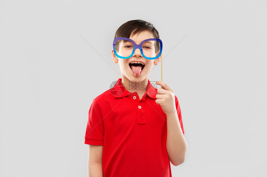 派道具,照片亭童的快乐的小男孩穿着红色马球T恤,戴着大纸眼镜,灰色背景上露出舌头快乐的男孩,露出图片