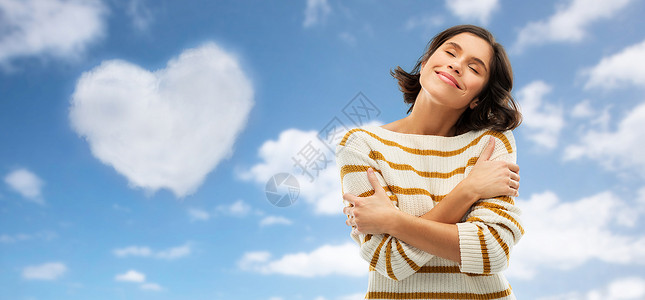 天空瓦伦丁情人节,爱的人快乐的轻女人穿着条纹套头衫,闭着眼睛拥抱自己蓝天云的形状的心脏背景女人心形的云上拥抱自己背景