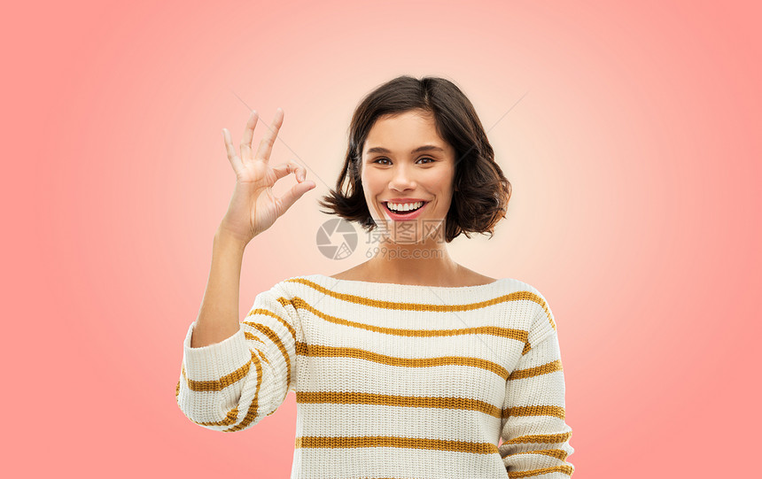 手势人的快乐的微笑轻女子穿条纹套衫,OK手标志活珊瑚粉红色的背景快乐微笑的女人穿着条纹套衫,表现得很好图片