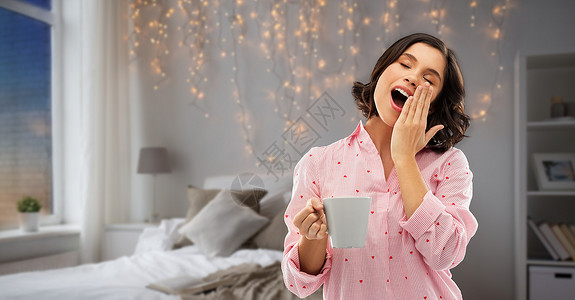 人们就寝时间的快乐困倦的轻妇女睡衣与咖啡打哈欠夜间卧室花环灯光背景快乐的哈欠轻的女人睡衣咖啡背景图片