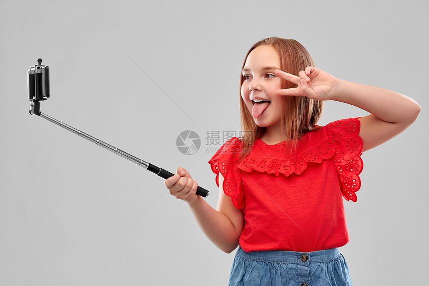 童人的美丽的微笑女孩穿着红色衬衫裙子,用智能手机自拍杆上拍照,并灰色背景上表现出平女孩用智能手机自拍杆上图片