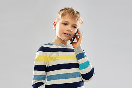 打电话小男孩技术,沟通人的小男孩穿着条纹套头衫,灰色背景下打电话给智能手机穿着条纹套头衫的男孩智能手机上打电话背景