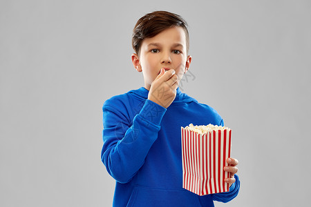 快餐,童人的男孩穿着蓝色帽衫,吃爆米花条纹纸桶灰色背景穿着蓝色帽衫的男孩吃爆米花背景