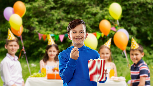 快餐,童人的快乐的微笑男孩穿着蓝色帽衫,夏季公园背景的生日聚会上,条纹纸桶上吃爆米花微笑的男孩生日聚会上吃爆背景