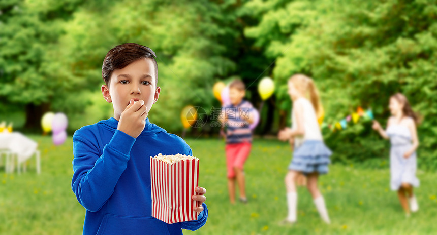 快餐,童人的男孩穿蓝色帽衫,吃爆米花条纹纸桶超过朋友生日聚会夏季公园背景男孩生日聚会上吃爆米花图片