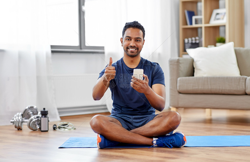 运动,技术健康的生活方式微笑的印度男人,智能手机坐运动垫上,家里竖大拇指印度男人家里用智能手机运动垫上图片