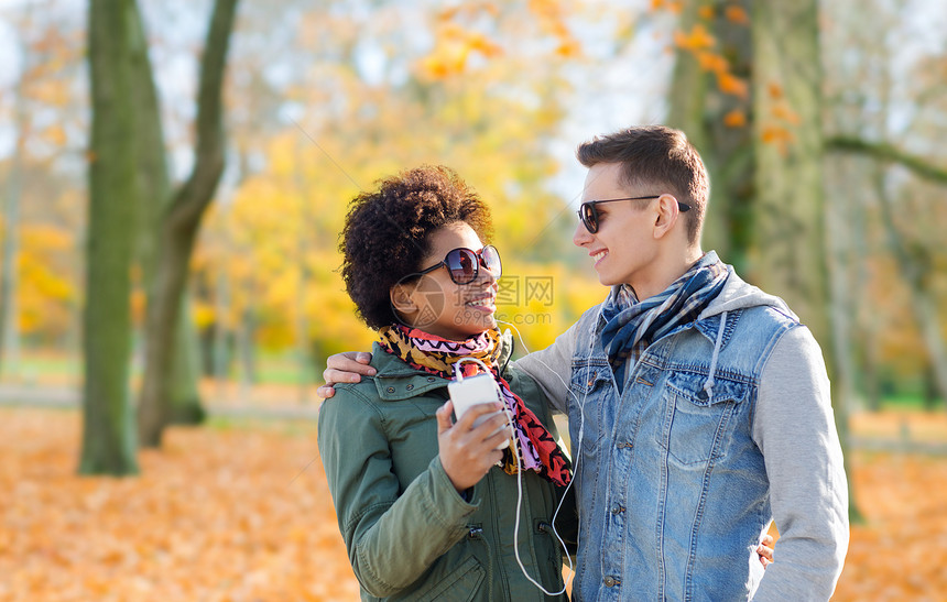 夏天,技术,人友谊的微笑混合种族夫妇与智能手机耳机听音乐秋天的公园背景秋天智能手机耳机图片