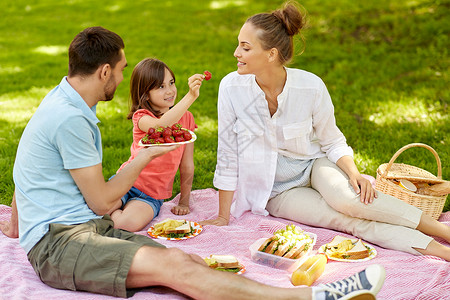 家庭休闲人的快乐的母亲父亲女儿夏季公园野餐吃草莓家人公园野餐时吃草莓图片