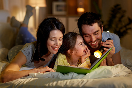 外国父母人们家庭的快乐的母亲,父亲小女儿家里晚上床上用手电筒看书快乐的家庭阅读书晚上床上家背景