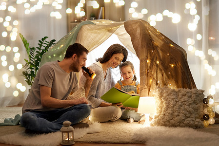 阅读灯家庭,潮格人的快乐的母亲,父亲小女儿阅读书与火炬灯孩子们的帐篷晚上家孩子们家帐篷里快乐的家庭读物背景