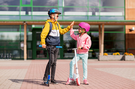 酷滑板车教育,童人的快乐的学校孩子戴着头盔,背包骑着滑板车,户外高五学校的孩子骑滑板车击掌背景
