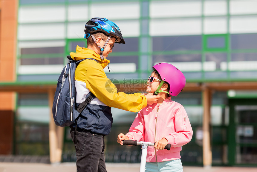 安全,童护理快乐的学校男孩与背包紧固女孩的头盔滑板车骑学校男孩为滑板车系紧女孩的头盔图片