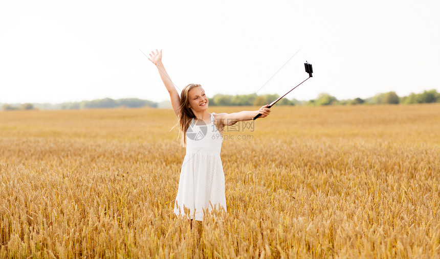 技术,夏天人们的快乐的轻女孩穿着白色连衣裙,用智能手机谷类食品领域的自拍杆上拍照快乐的轻女孩用智能手机自拍图片