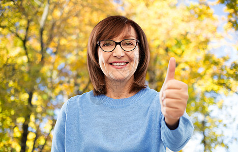 手势,视觉老人的微笑的老年女子眼镜上的肖像,秋天的公园背景上竖大拇指老妇女的肖像竖大拇指背景图片