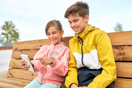 府前街童技术人的快乐的孩子兄弟姐妹智能手机坐户外的木街长凳上孩子们着智能手机坐长凳上背景