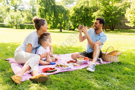给你孩子买房了嘛家庭休闲人的父亲夏季公园野餐时用智能手机拍下了幸福的母亲两个小儿子的照片父亲公园野餐时给家人拍照背景