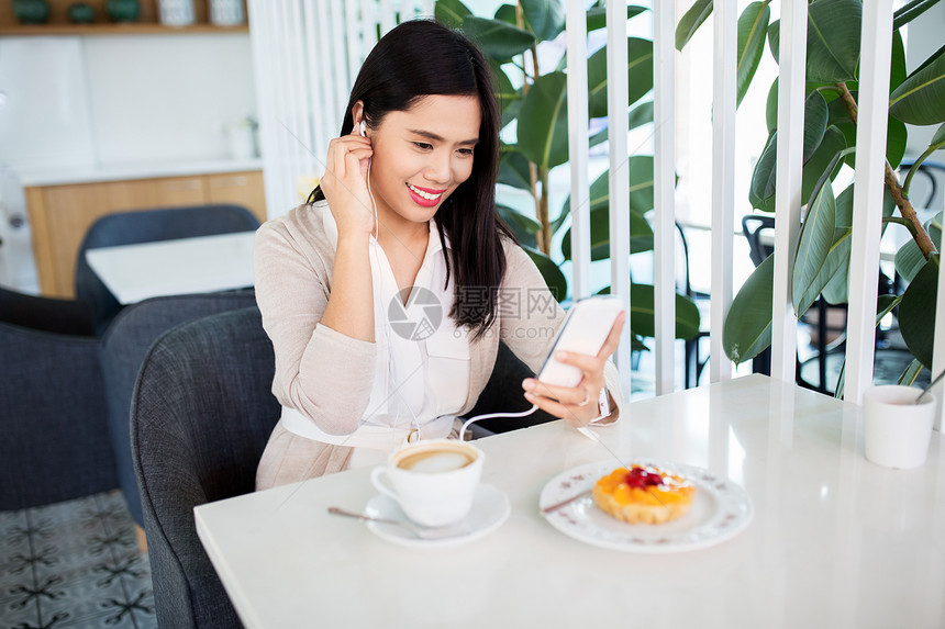 技术人的快乐的亚洲妇女与智能手机,耳机蛋糕听音乐咖啡馆咖啡店咖啡馆智能手机耳机的亚洲女人图片
