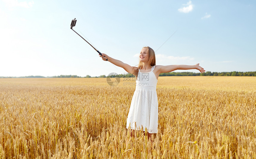 技术,夏天人们的快乐的轻女孩穿着白色连衣裙,用智能手机谷类食品领域的自拍杆上拍照快乐的轻女孩用智能手机自拍图片