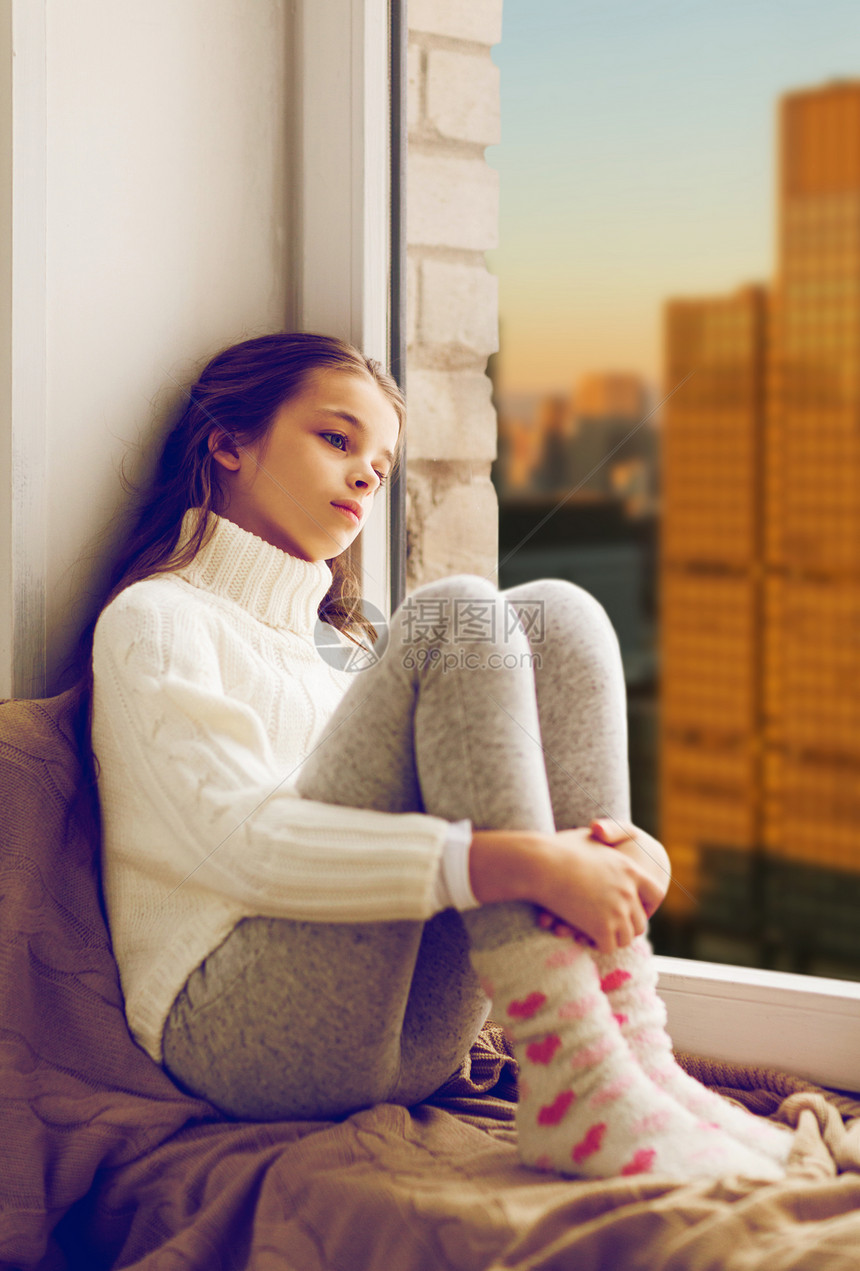 童,悲伤人的悲伤的美丽女孩穿着毛衣,坐家里的窗台上,城市的摩天大楼背景上悲伤的女孩坐窗台上,城市的窗户上图片