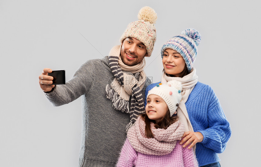 家庭,诞节技术快乐的母亲,父亲小女儿穿着冬天的衣服,用智能手机灰色背景下自拍幸福的家庭用智能手机自拍图片