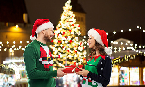 幸福的夫妇戴着诞老人帽子并穿着绿色的毛衣诞市场的灯光背景前赠送礼品盒图片