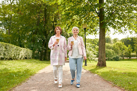 桃花散步喝饮料老退休人的两个老妇女朋友喝咖啡沿着夏季公园散步老年女子朋友公园喝咖啡背景