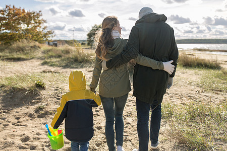 后方的家庭家庭,休闲人的快乐的母亲,父亲小儿子带着沙子玩具套件沿着秋天的海滩散步幸福的家庭沿着秋天的海滩散步背景
