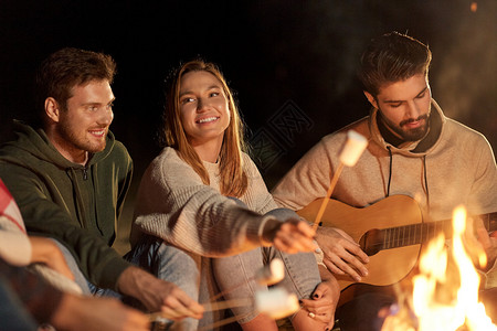 休闲人的群微笑的朋友坐篝火旁,烤棉花糖,晚上弹吉他朋友们烤棉花糖弹吉他图片