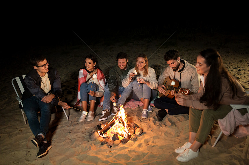 休闲人的群微笑的朋友坐海滩上的篝火旁,晚上烤棉花糖朋友们海滩上篝火上烤棉花糖图片