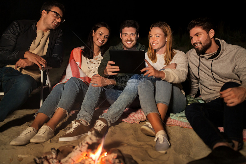 休闲人的群微笑的朋友坐营地火灾与平板电脑电脑海滩上的夜晚朋友与平板电脑海滩火灾晚上图片