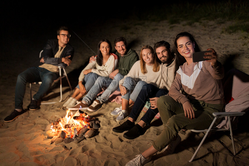 休闲人的群微笑的朋友坐篝火上,海滩上用智能手机自拍快乐的朋友海滩的篝火上自拍图片