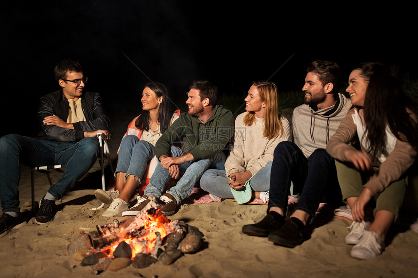休闲人的群微笑的朋友晚上坐海滩上的篝火旁群朋友坐海滩上的篝火旁图片