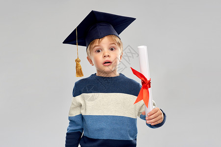灰色毕业帽子学校,教育学快乐的小男孩戴着学士帽灰背景文凭的黑板带着文凭的灰泥板上的小男孩背景