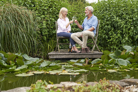 成熟的夫妇湖边的木制码头上坐椅子上用香槟庆祝图片