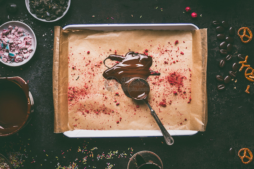 勺子与融化的巧克力托盘上与红色干果粉末顶部黑暗的厨房桌子背景与各种调味成分的巧克力棒制作,顶部视图清洁食品的图片