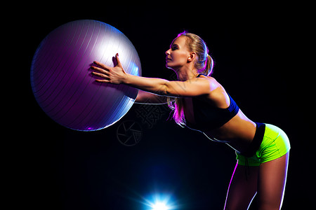 轻的运动女训练与健身球黑色背景与蓝色耀斑灯健身球的女人图片