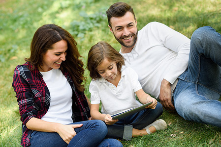 城市公园玩平板电脑的幸福家庭爸爸妈妈小女儿坐草地上笑图片