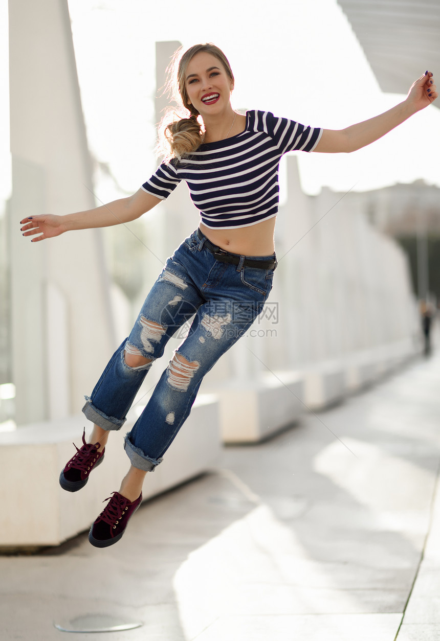趣的金发女人城市背景下跳跃漂亮的轻女孩穿着条纹T恤蓝色牛仔裤街上漂亮的俄罗斯女,辫子图片
