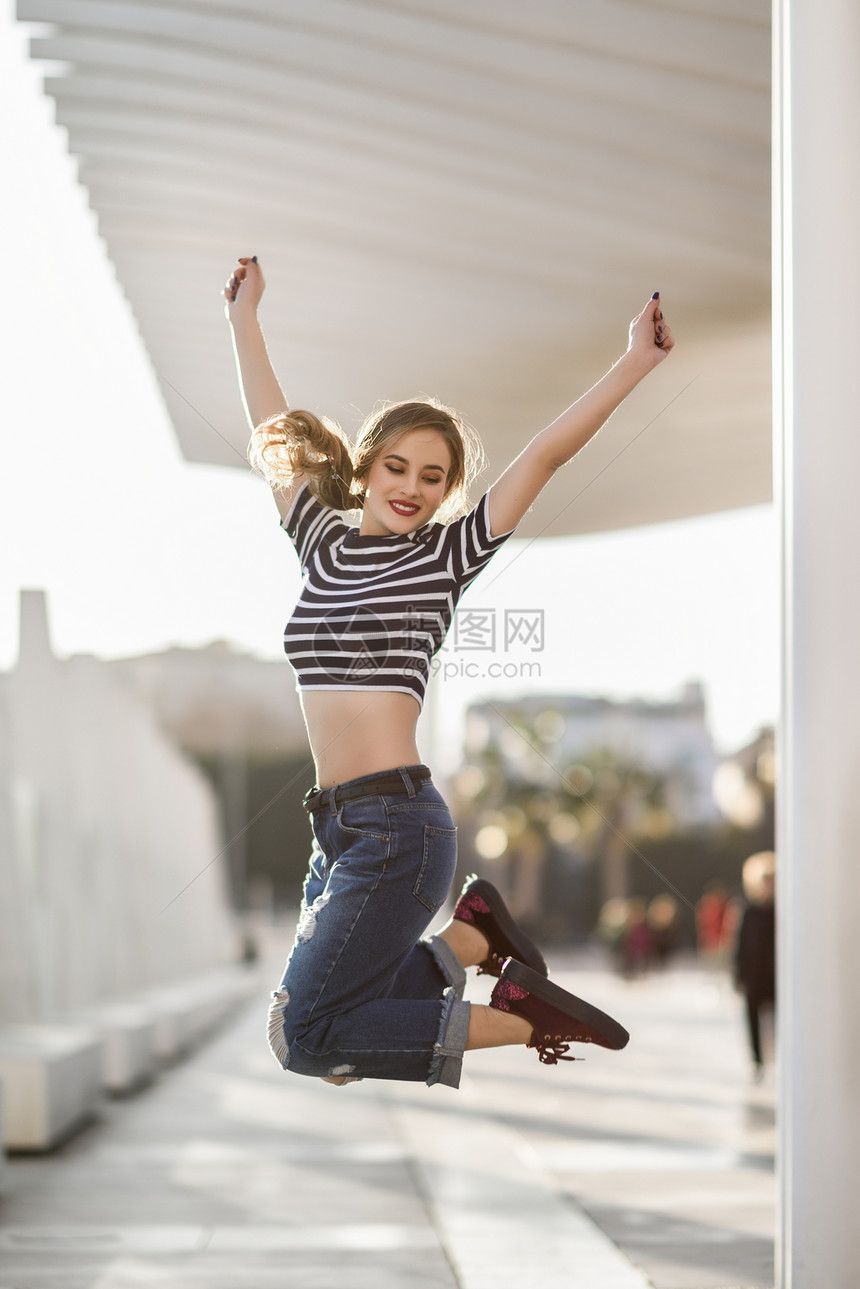 趣的金发女人城市背景下跳跃漂亮的轻女孩穿着条纹T恤蓝色牛仔裤街上漂亮的俄罗斯女,辫子图片