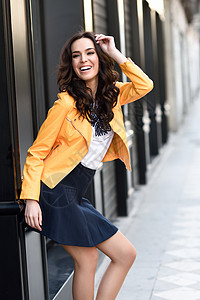 轻的黑发女人,时尚模特,穿着橙色现代夹克蓝色裙子,城市背景下大笑图片